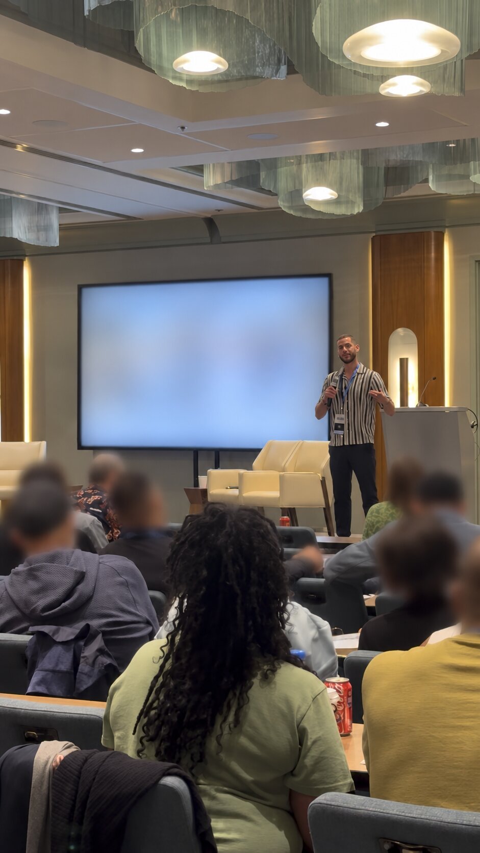 Facilitator speaking to a seated group, audience and presentation screen blurred for privacy
